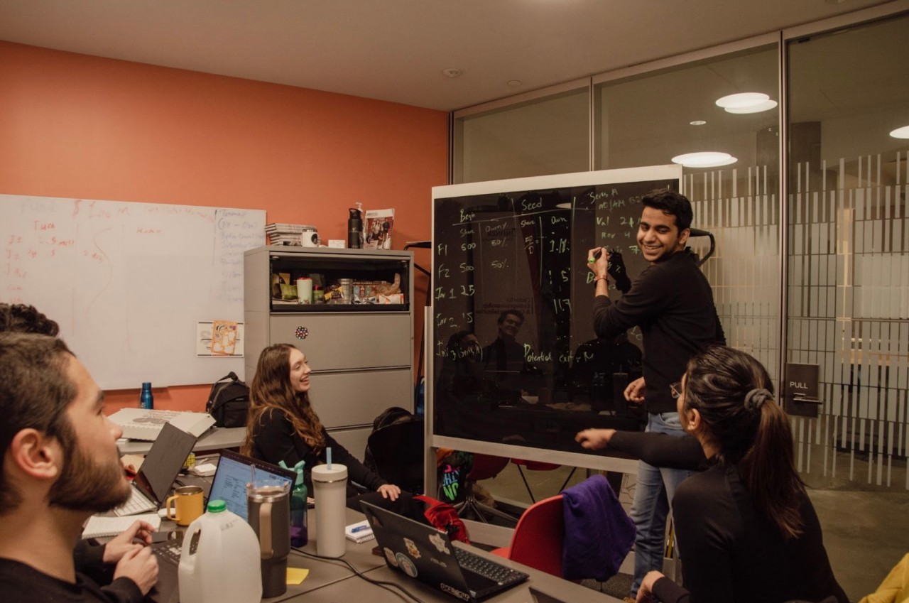 Students gather in a conference room and discuss ideas as one person writes on a board.
