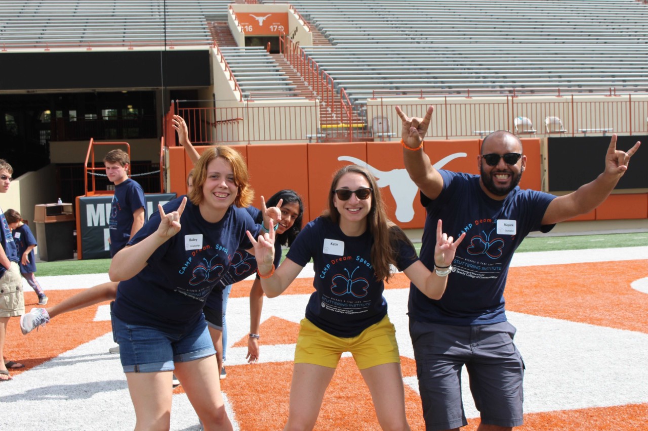 Three people on a field make a University of Texas gesture with their hands.
