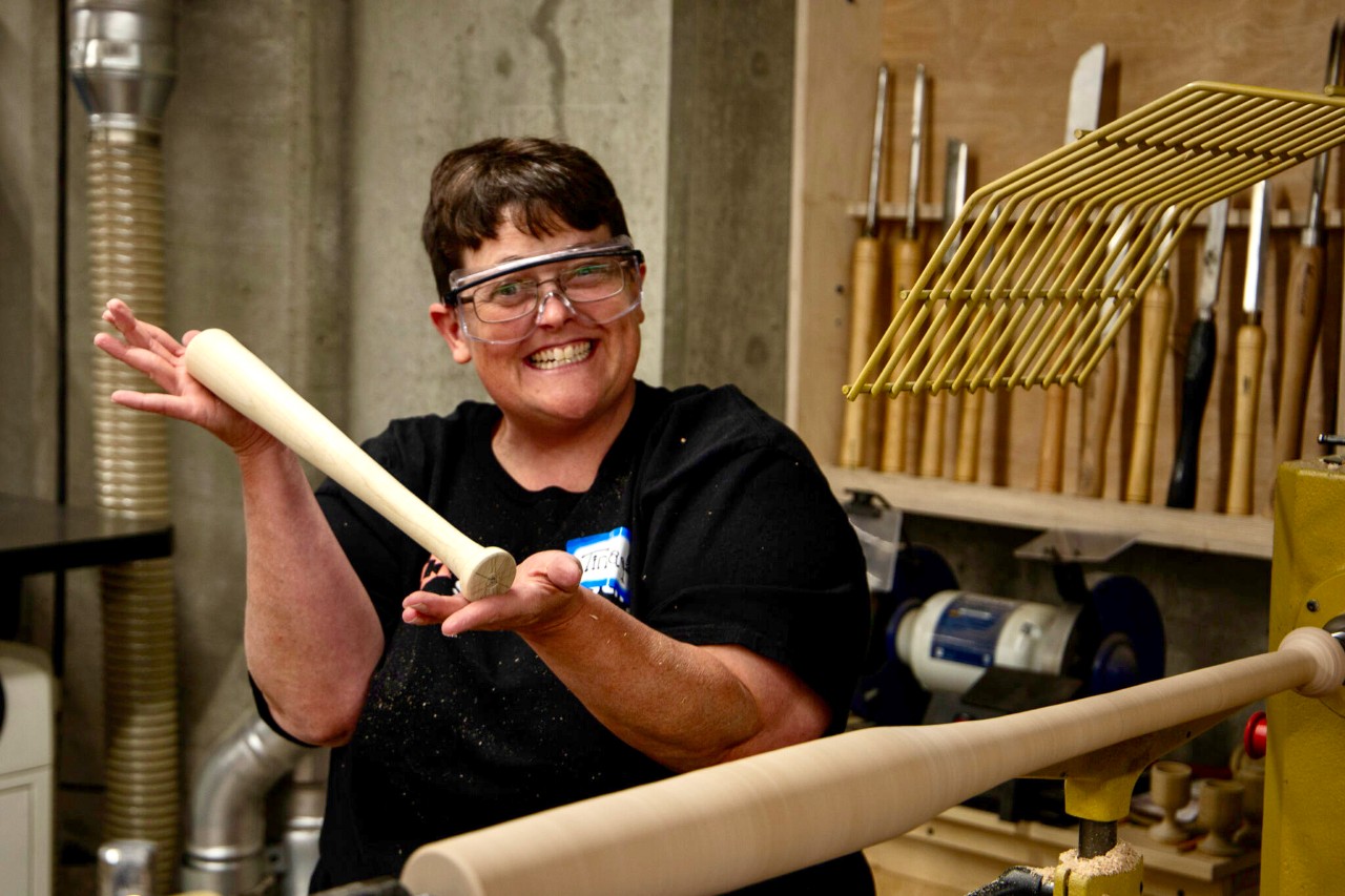A smiling woman in a makerspace holds a wooden baseball bat near a woodworking machine.
