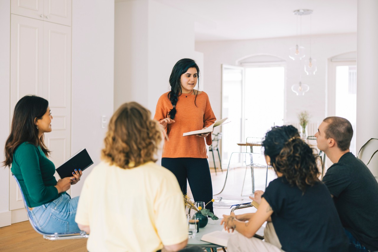 adult woman standing reading aloud from a book to other adults 