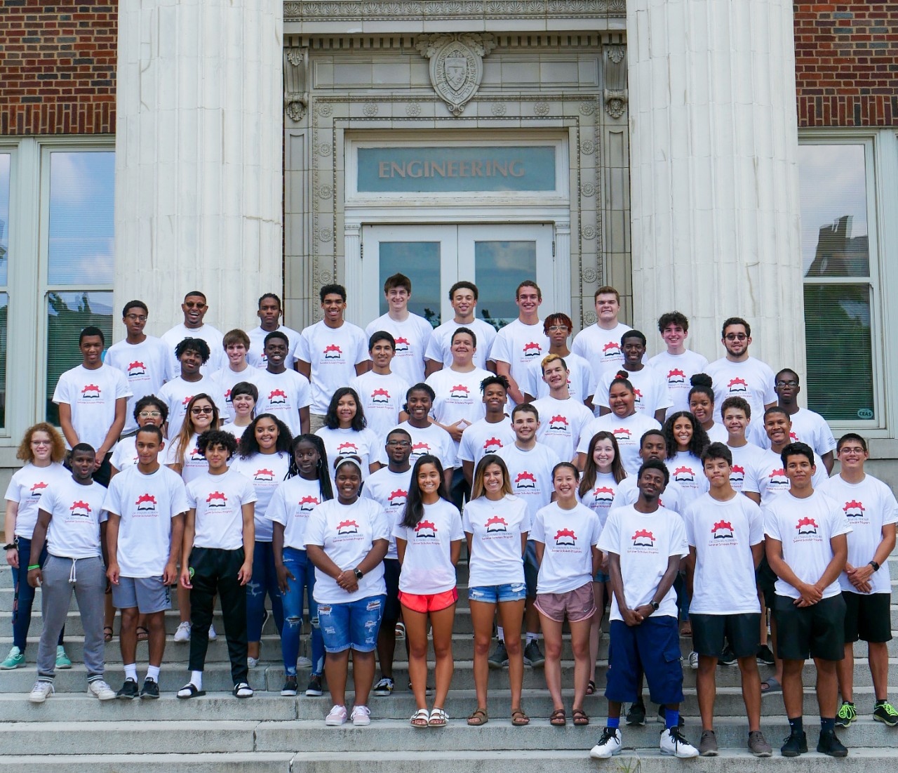 A group of students stand on the Baldwin steps at the University of Cincinnati. They wear matching white t-shirts.