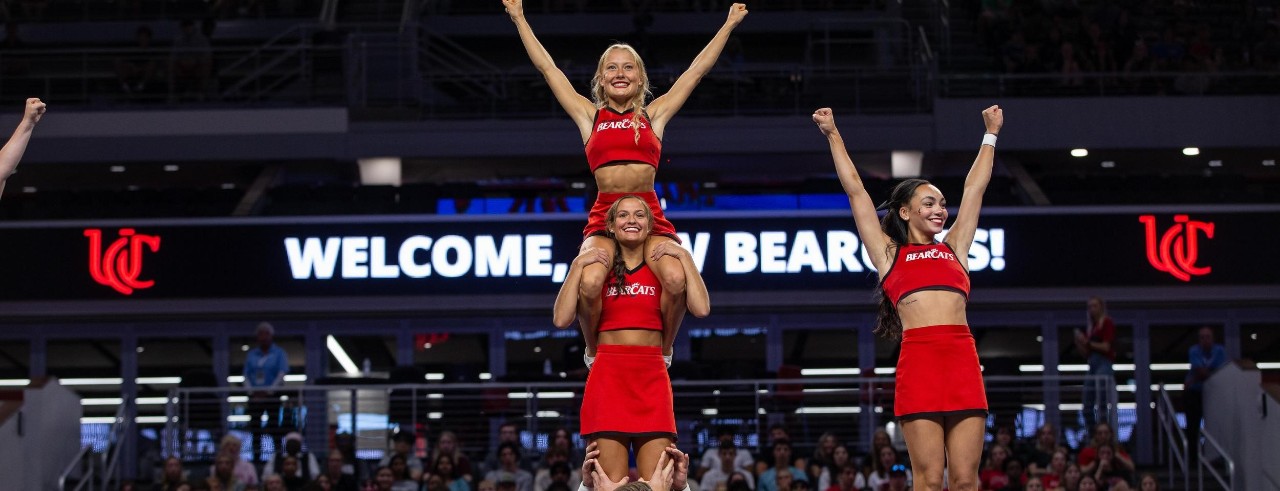 male members of cheer team hold three female cheer team members up in the air with a welcome bearcat sign in the background