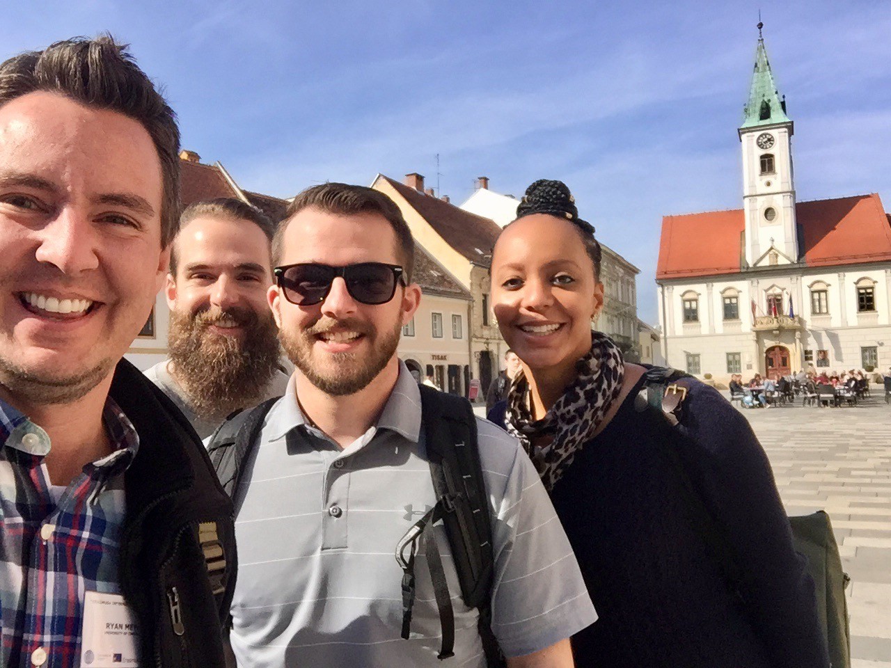 Four people pose for a selfie in Varaždin, with municipal buildings and shops lining the plaza behind them