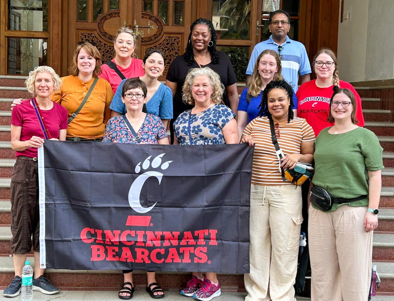A group holding a banner that reads “Cincinnati Bearcats” poses on the steps in front of a building with ornately carved doors.