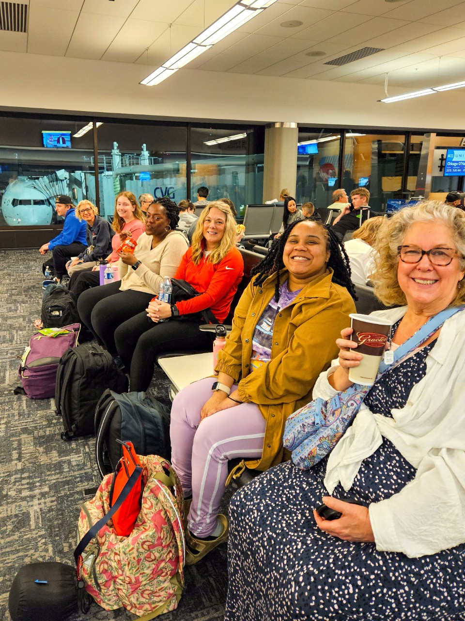 People sit in a row of chairs at the Cincinnati airport, smiling toward the camera. One holds a large cup of tea, and luggage is tucked around their feet.
