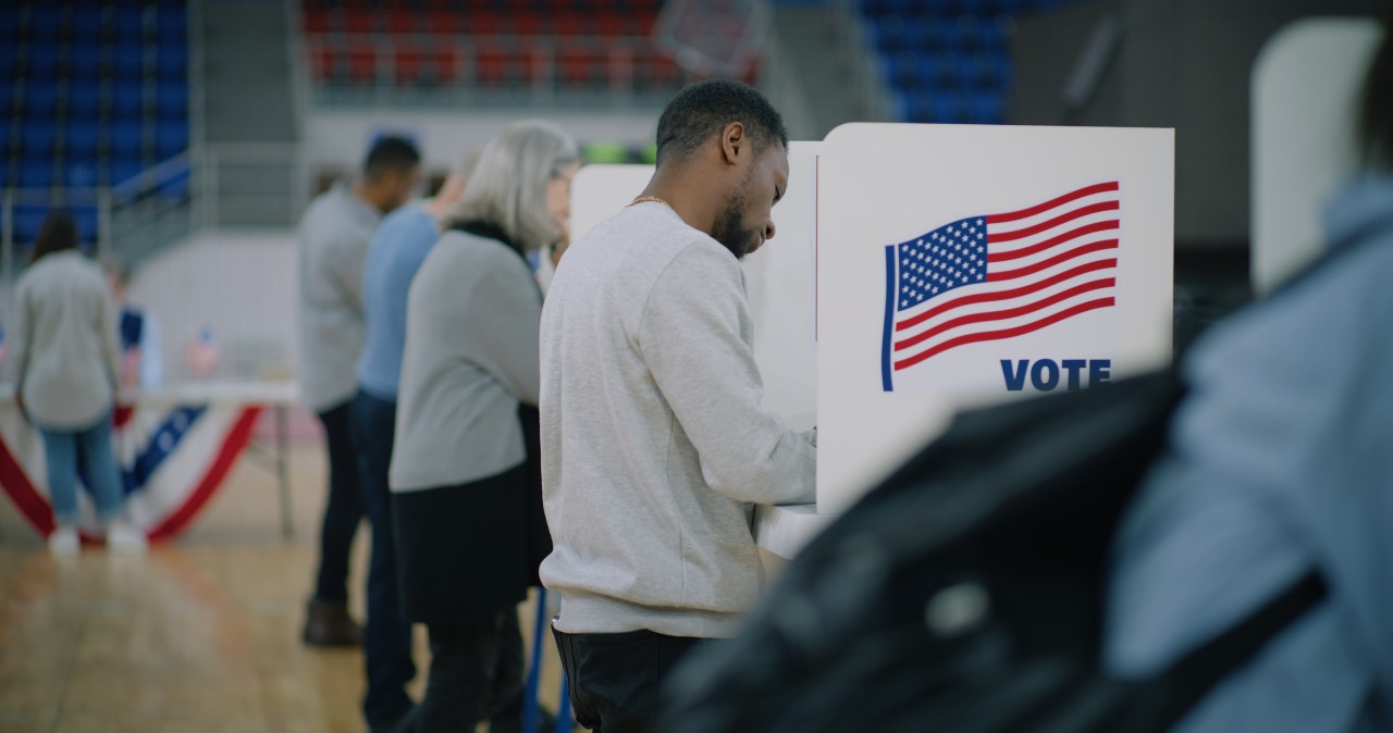 People standing at voting booths, while man approaches with voting bulletin in hand