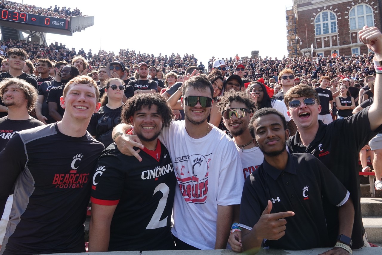 six young men in UC swag dominant the image with hundreds of fans cheering behind them in Nippert Stadium
