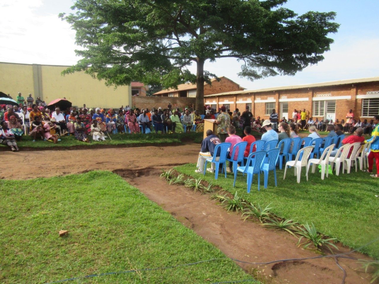 People sit on either side of a podium, one person stands at the podium speaking. The meeting is held outside