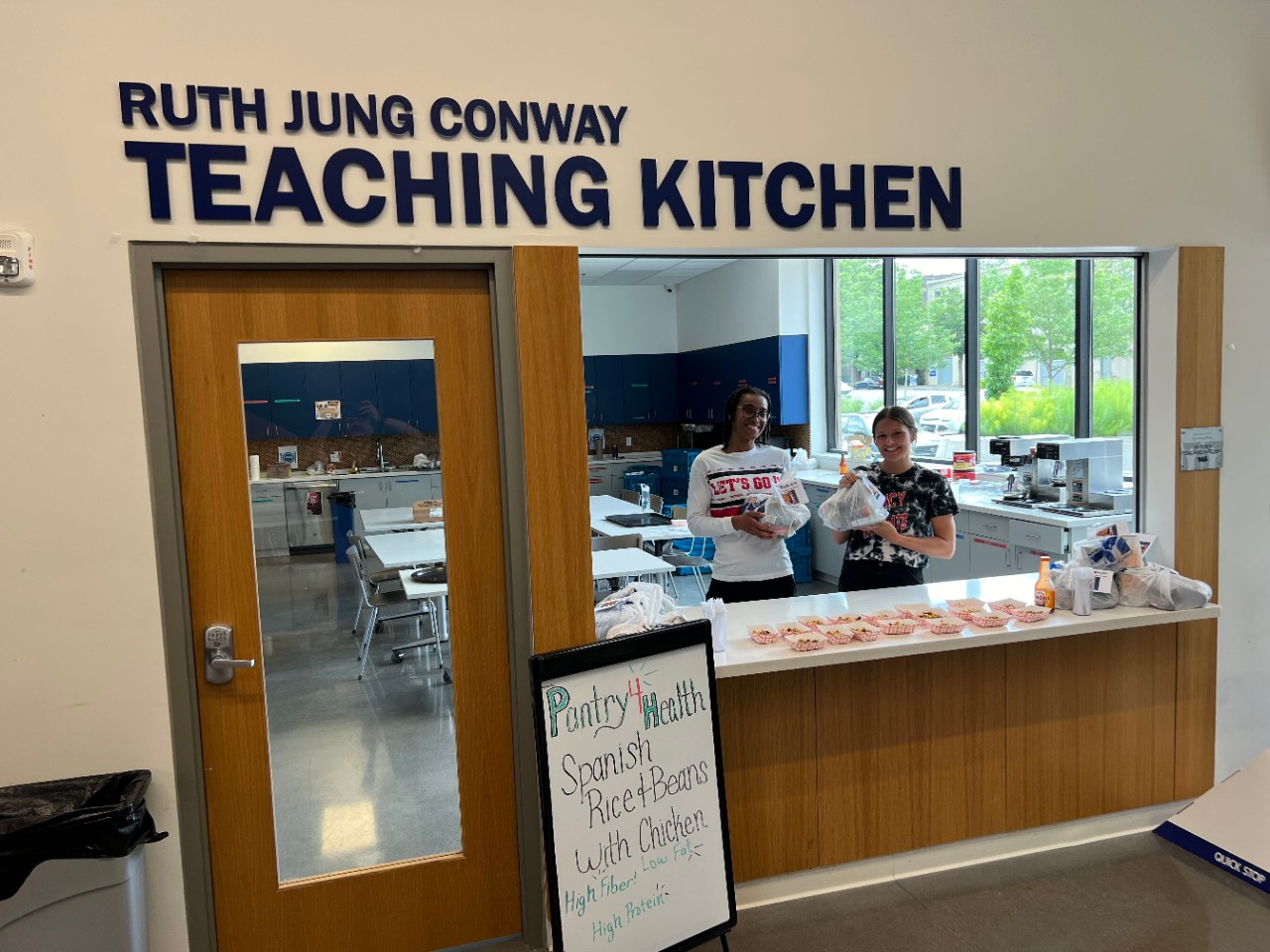 Two students hold up bags of food with samples of food on a counter in front of them.