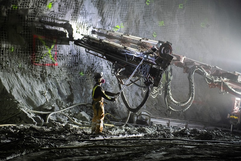 A worker in protective gear uses heavy equipment on a cavern wall illuminated with construction lights.