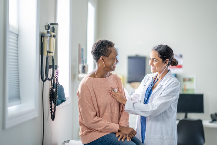 A woman sits on an exam table as her female doctor listens to her heart.