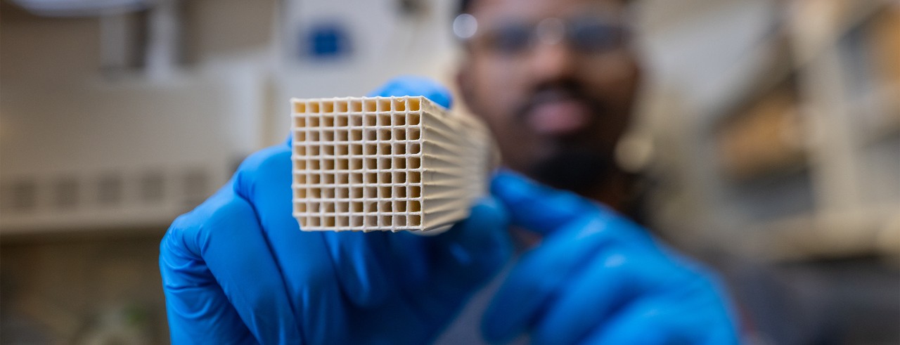 A UC engineering student holds up a honeycombed block with two gloved hands in an engineering lab.