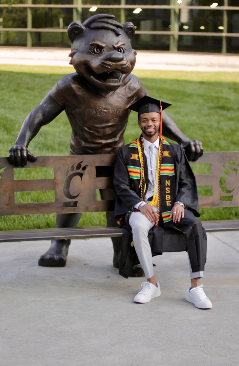 Michael Hunter sits on a bench next to a statue of the University of Cincinnati Bearcat Mascot and wears his graduation cap and gown.