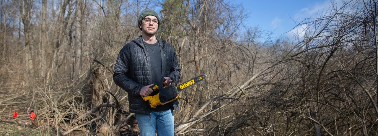 Griffin Paul holds a chainsaw in a patch of woods full of honeysuckle.