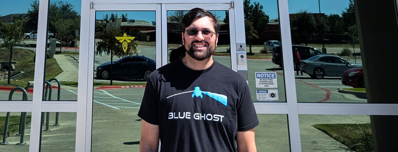 Ilyas Malik smiling in front of glass doors with Firefly Aerospace logo visible. He is wearing a Blue Ghost t-shirt
