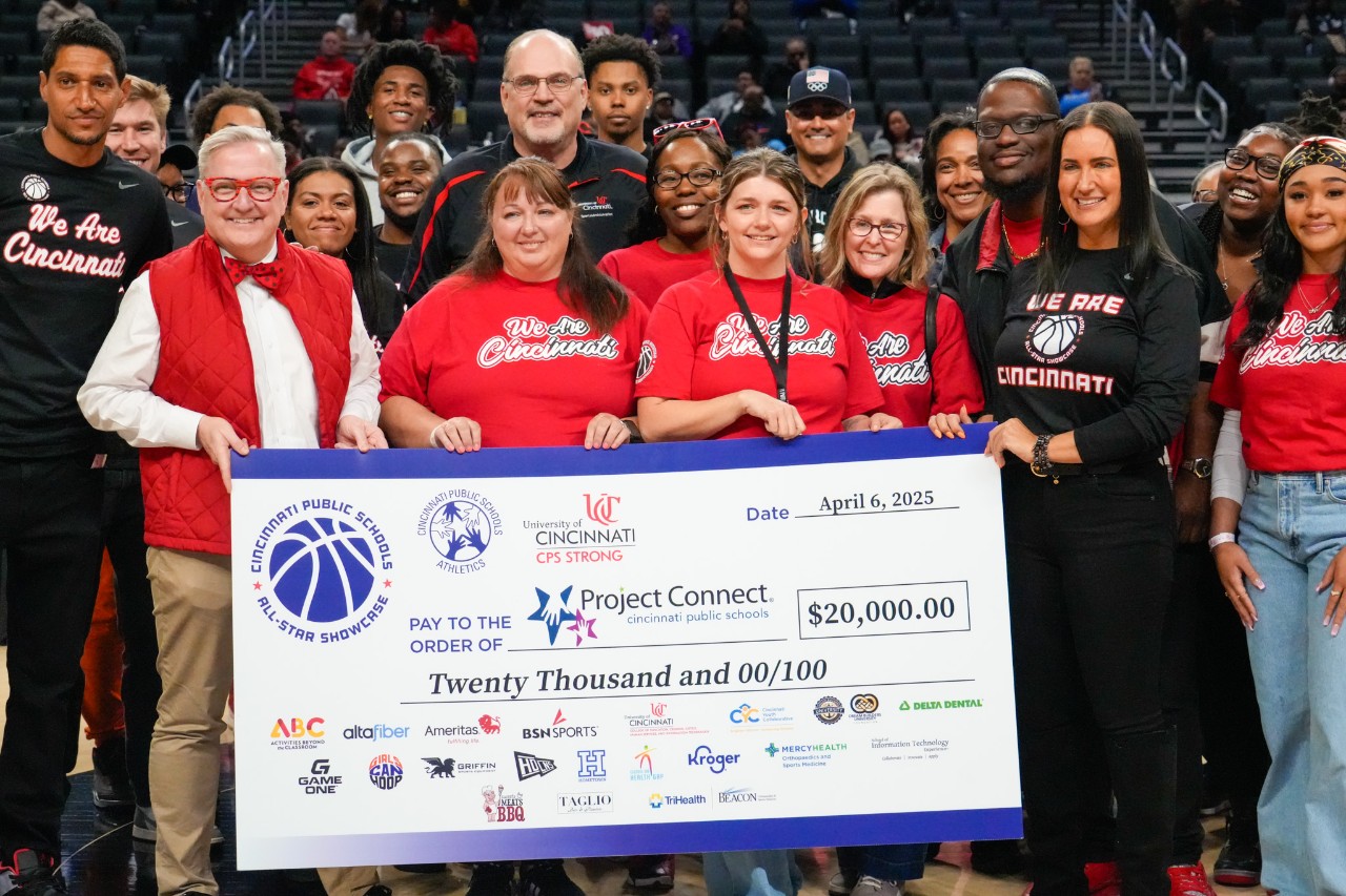 large crowd of Cincinnati Public Schools and UC official stand behind big poster board check of $20,000 on the basketball court 