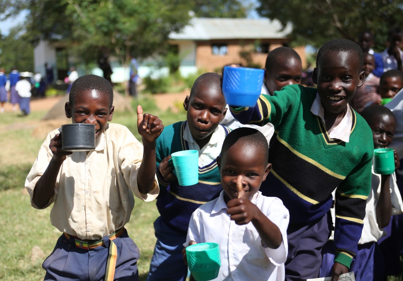Several Tanzanian children stand together holding plastic mugs of corn porridge.  