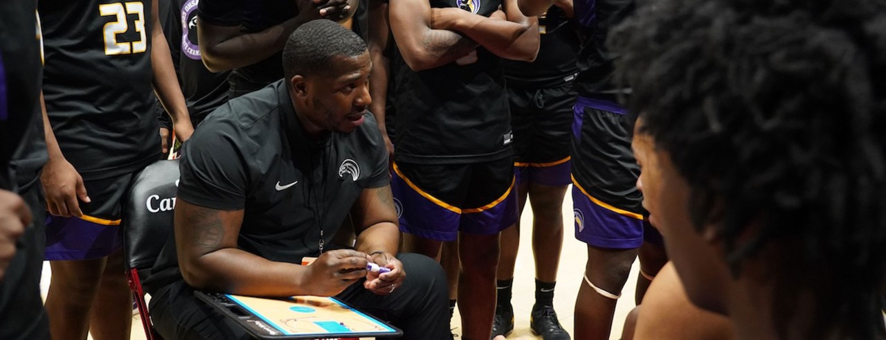 Aiken coach Derrell Black sits in a chair with a clipboard explaining a play to the Aiken Falcons varsity men's basketball team.