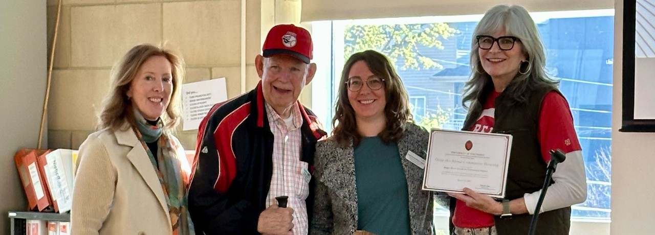 Four individuals smiling, with one holding a certificate. Two wear University of Cincinnati apparel. Indoors with windows in the background.