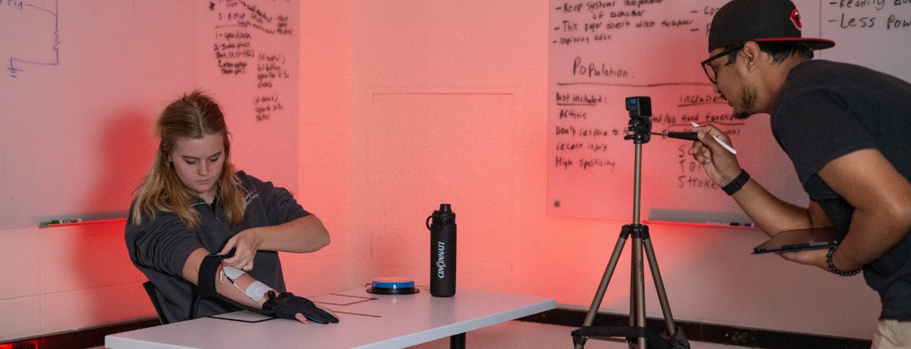 One graduate student sits at a table adjusting an exoskeleton prototype while another student stands behind a tripod with a camera on it taking pictures