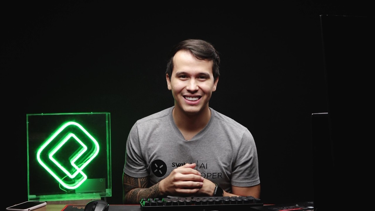 Adonai Vera Gonzalez sits at a desk with a black background and a green neon light displaying an arrow to his left. 