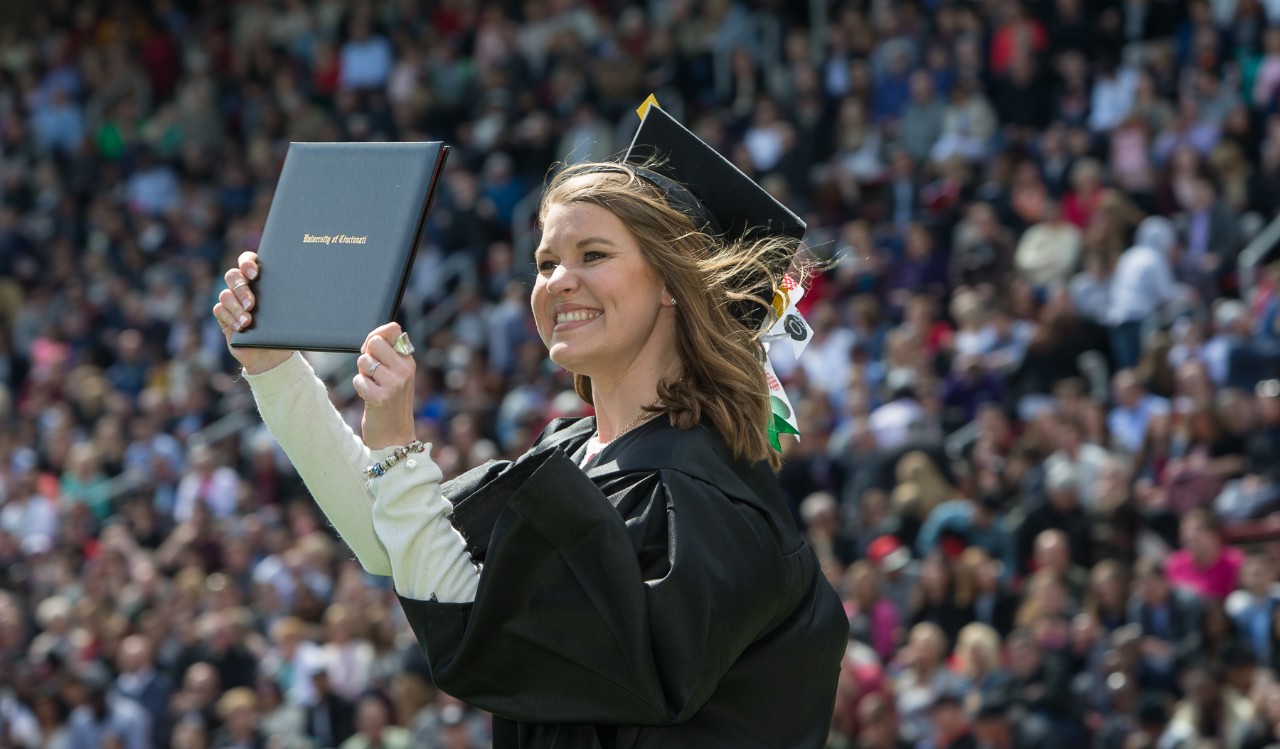 A graduate in cap and gown holds up her diploma cover with a crowd of people in the background at Nippert Stadium.
