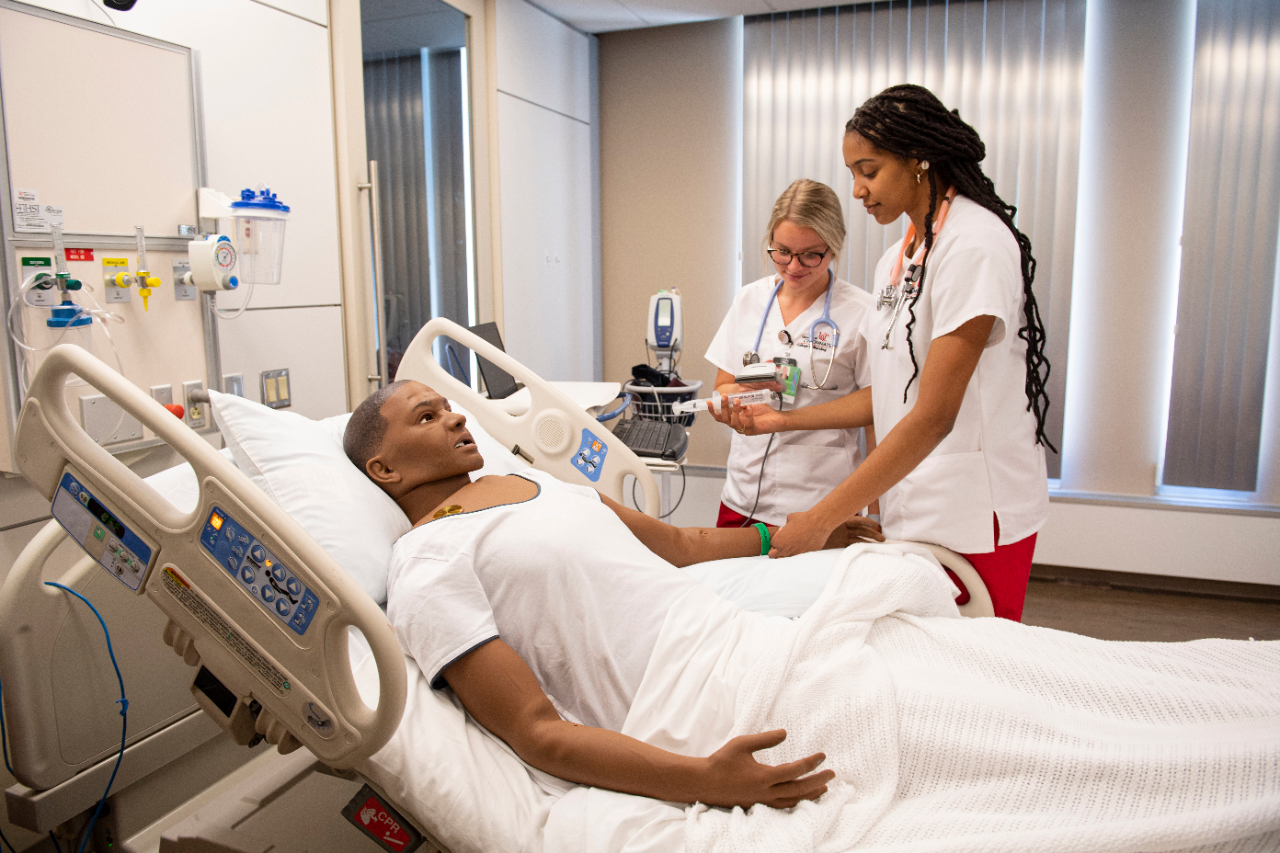 Two UC nursing students studying a dummy doll in a hospital bed for their future career