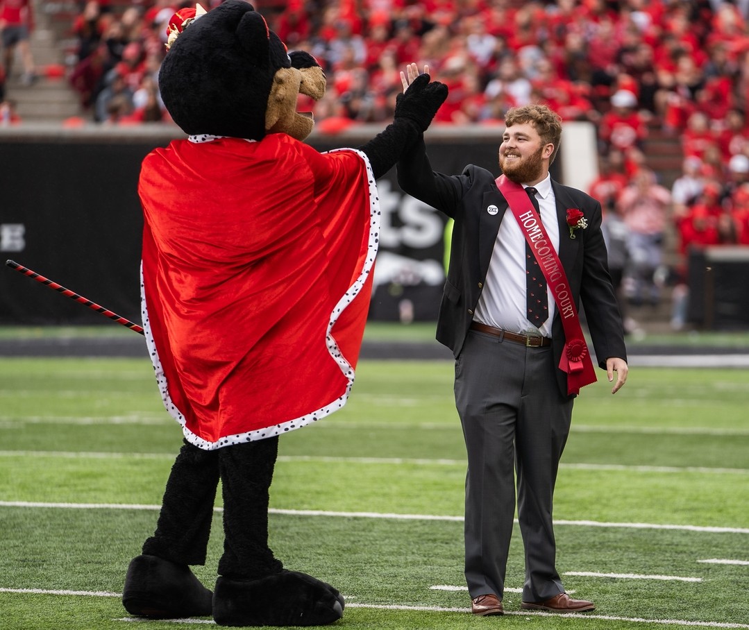 The Bearcat mascot high fives Evan Phelps, dressed in a suit and tie, on the Nippert Stadium turf.