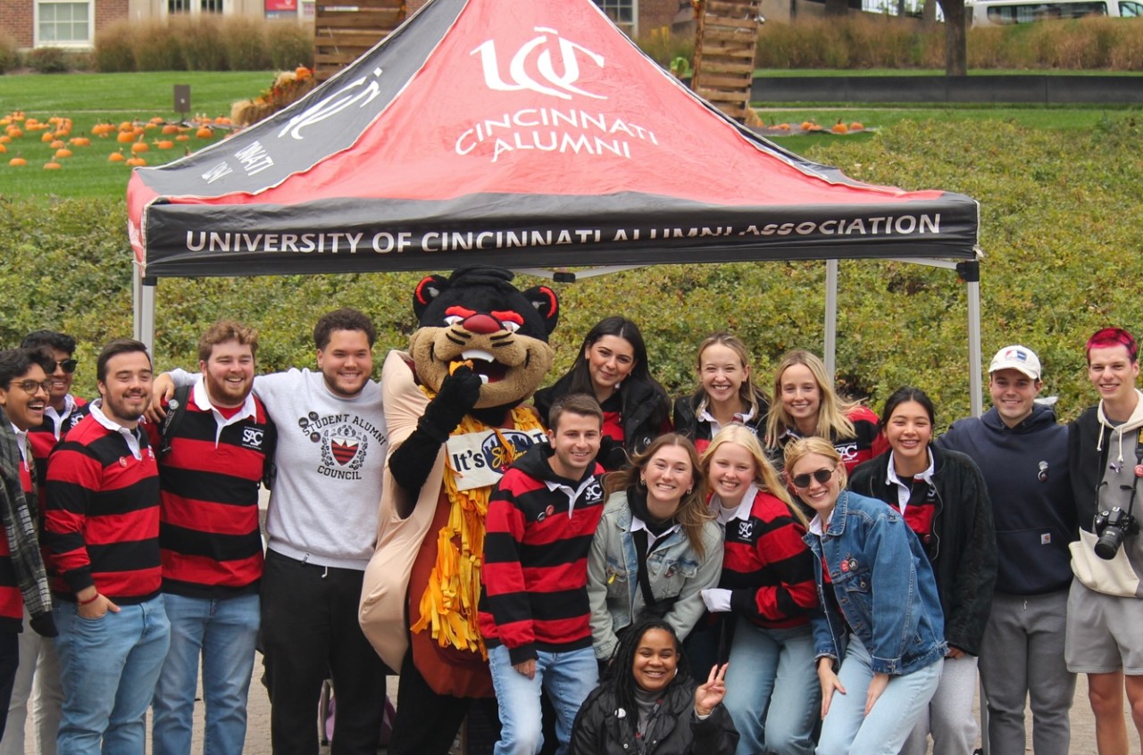 Students, some in red and black striped shirts, stand under a red and black tent and pose for a group photo.