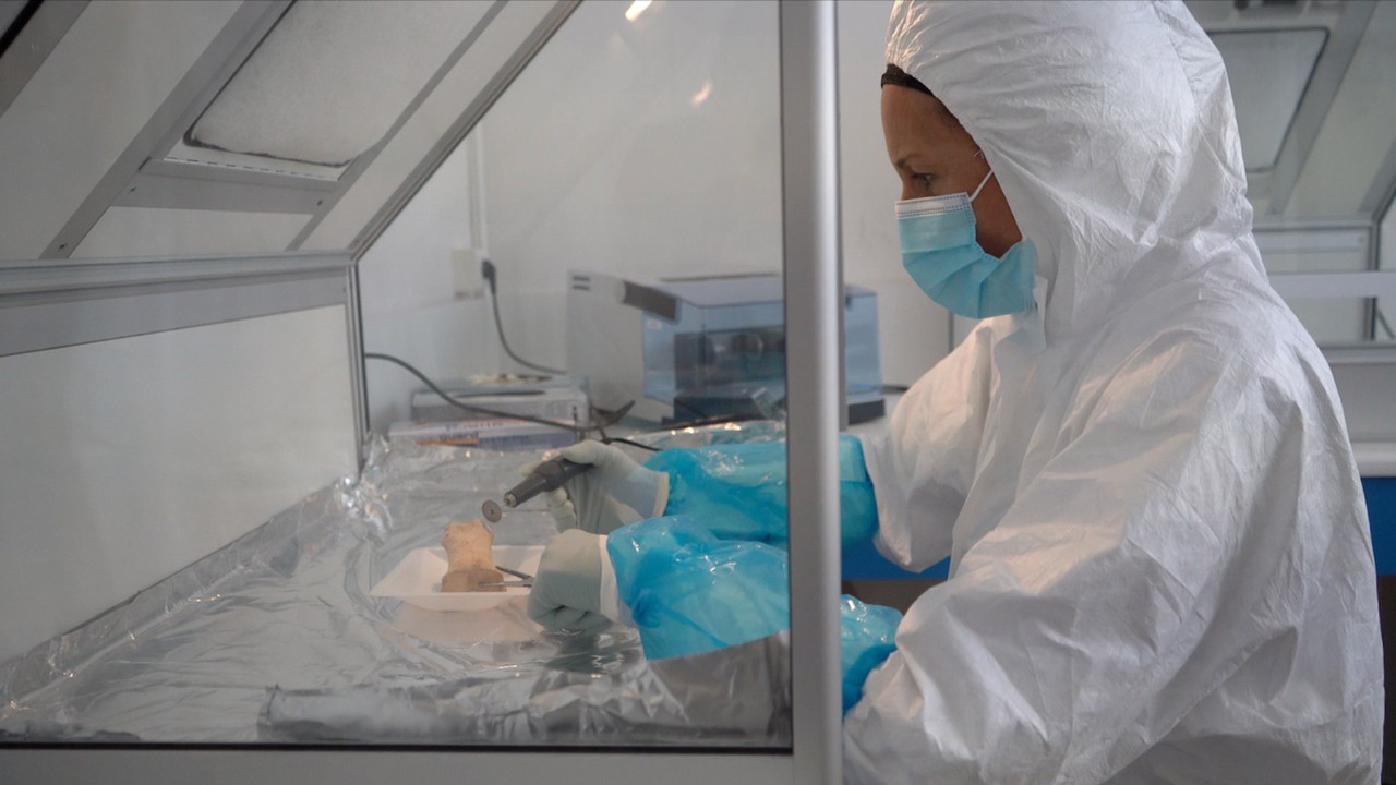 A scientist in protective clothing takes a sample on a lab bench.