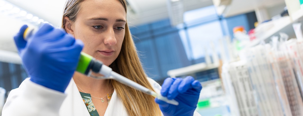 Katelyn Jansen, wearing a white lab coat and gloves, pipettes a sample in the lab