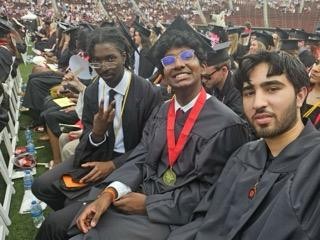 Sai Gollamudi shown sitting between two other students, all in cap and gown, during UC Commencement
