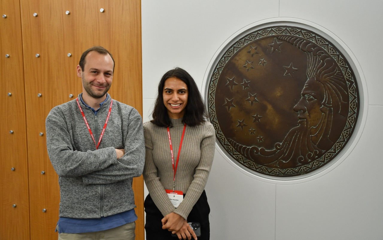 Professor Pietro Strobbia and chemistry student Manisha Sheokand stand in front of a bronze plaque of the P&G logo. 