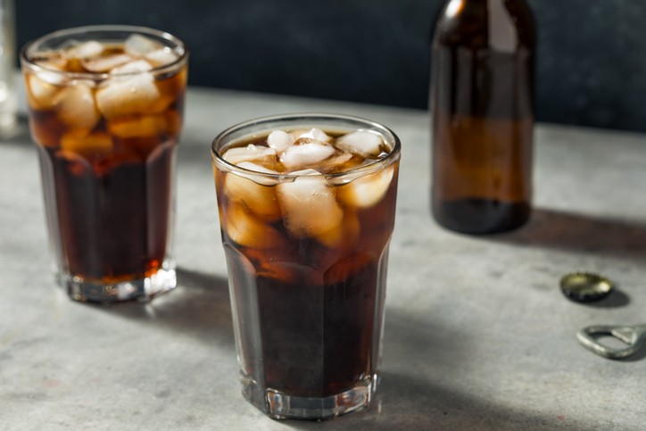 Two glasses of diet cola with ice on a counter in front of a brown glass bottle