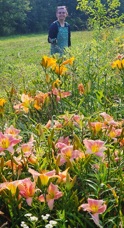 Person standing in a flower field smiling with lush greenery and bright orange flowers in the foreground.
