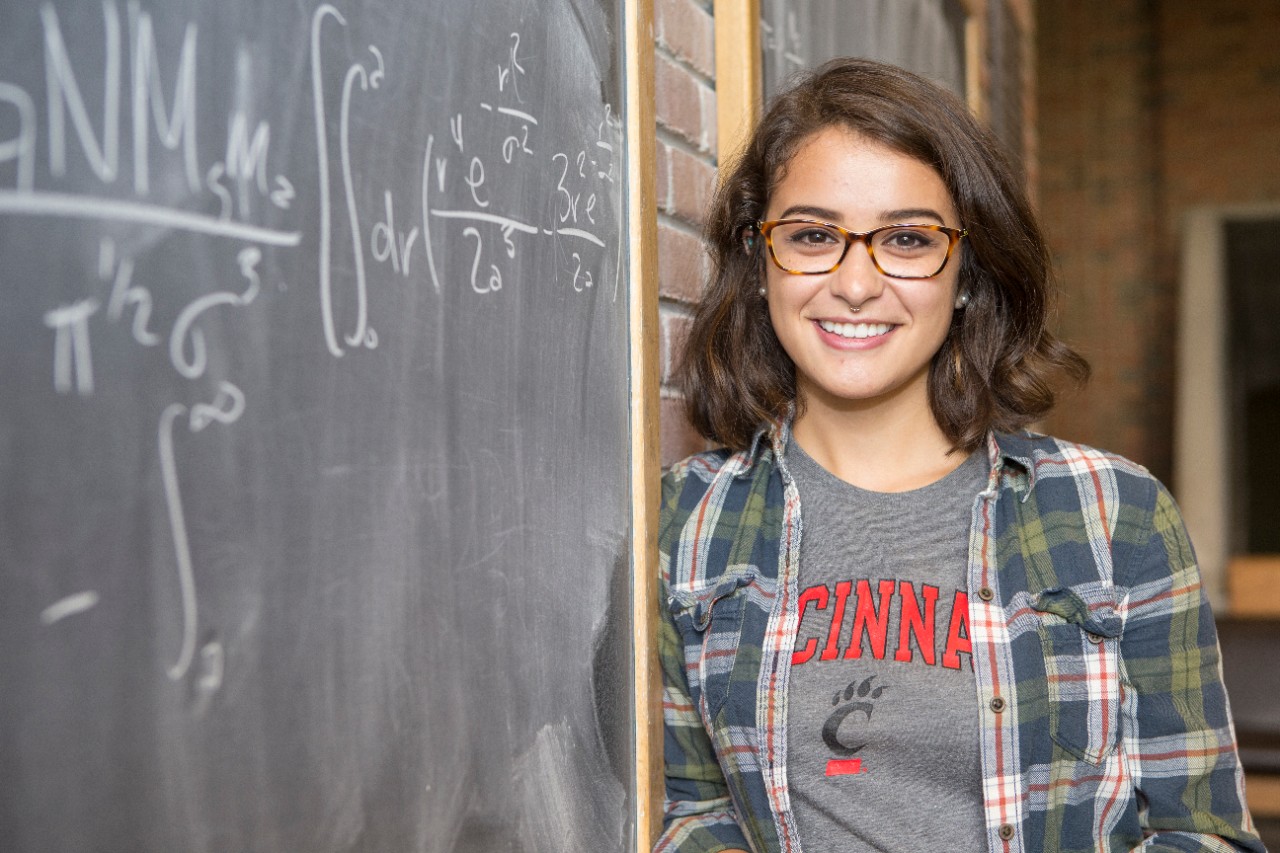 UC student wearing a UC shirt smiling for a photo next to a chalkboard