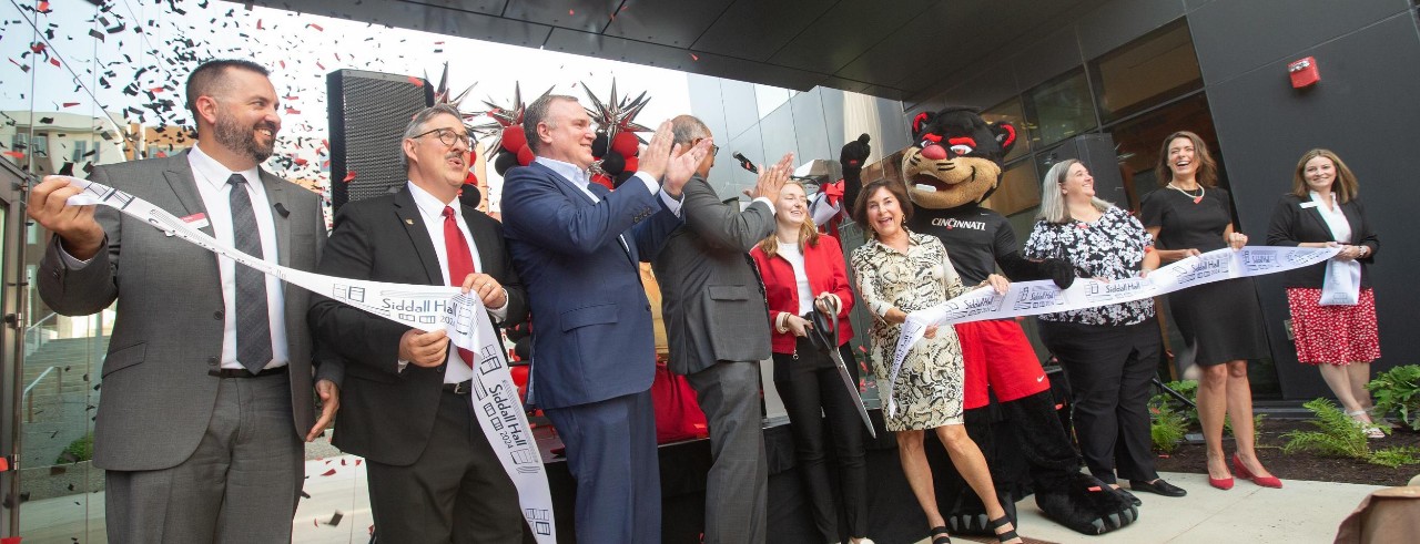 Several UC officials are shown holding a cut ribbon and celebrating Siddall Hall's grand reopening