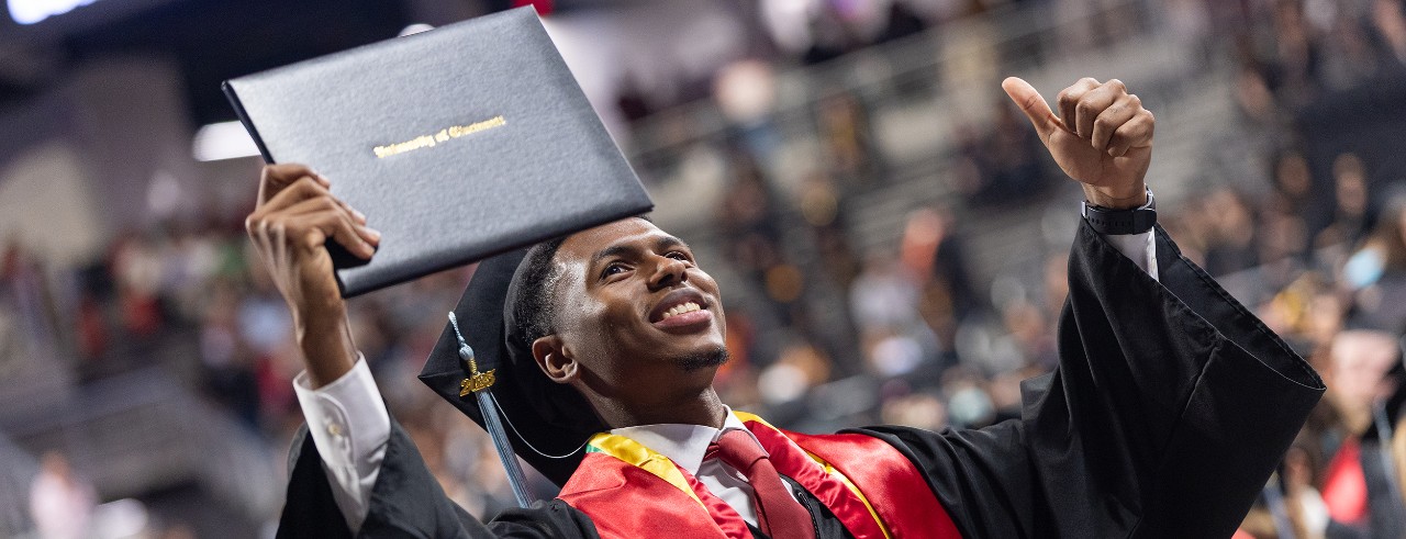 A UC grad in a cap and gown holds his diploma up triumphantly at Fifth Third Arena.