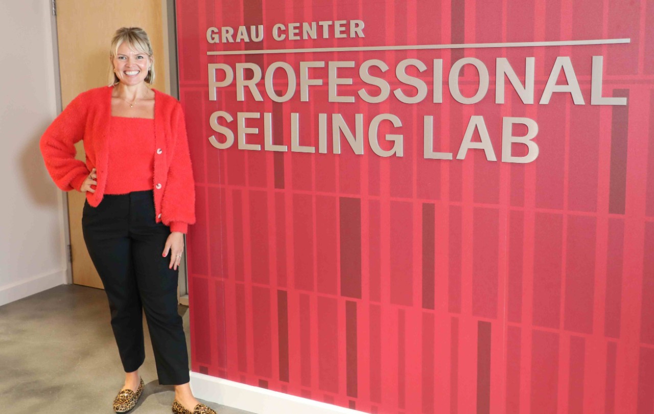A woman in a red top and black pants stands in front of a red and gray wall.