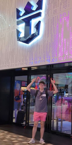 Price standing in front of a brightly lit Royal Caribbean logo inside what appears to be an onboard clothing store. Price is raising their hands to point at the logo and he is wearing a University of Cincinnati t-shirt.