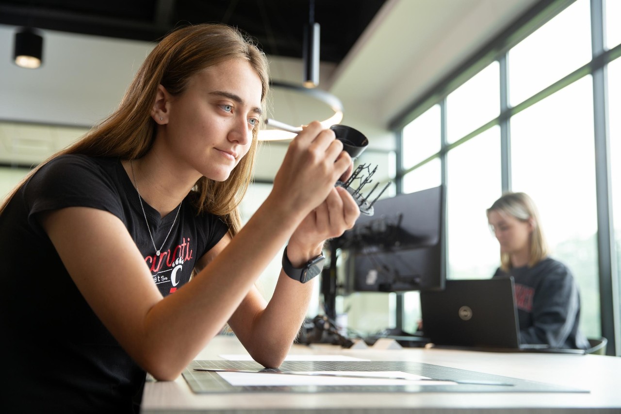 Brooke  Boeding shown sitting at work desk with mechanical instrument in hand and female student on a laptop is shown in the background