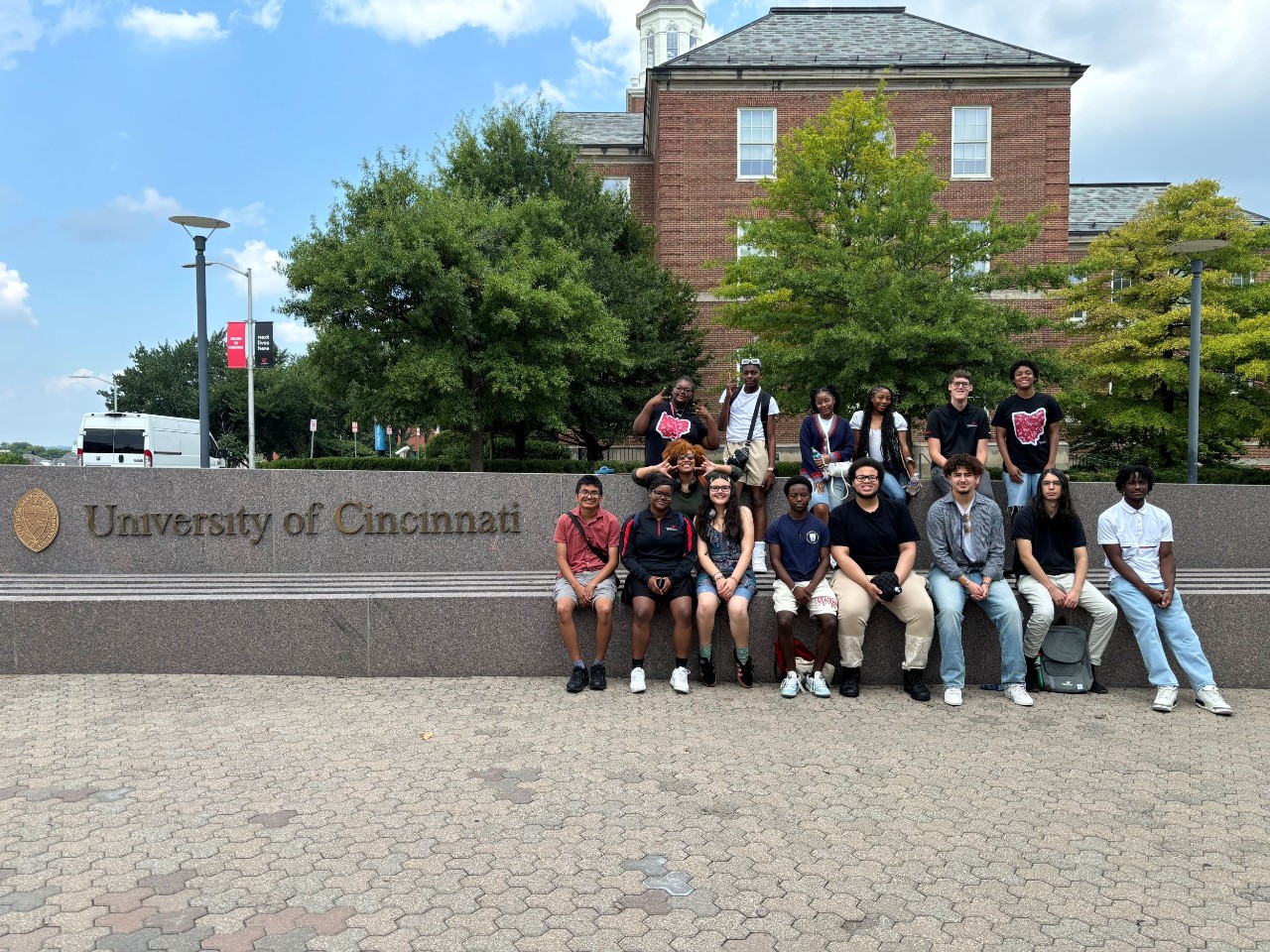 About 20 students sit next to the University of Cincinnati sign with the College of Arts & Sciences in the background