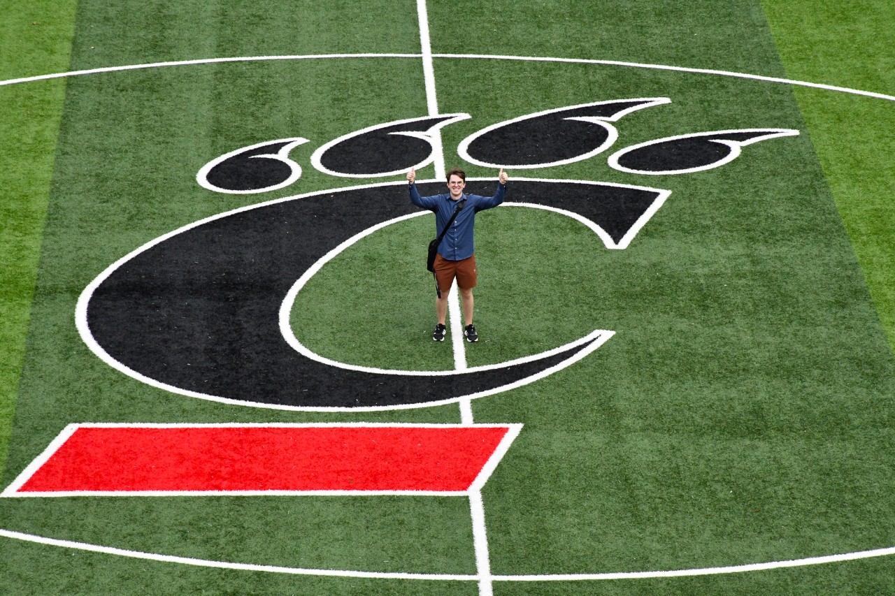 Samuel Hall holds his arms up on a football field decorated with UC's logo.