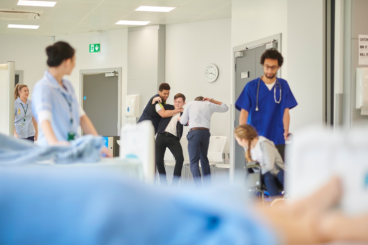 Image shows a security guard helping to subdue a visitor in an emergency room setting.  Healthcare workers stand nearby.