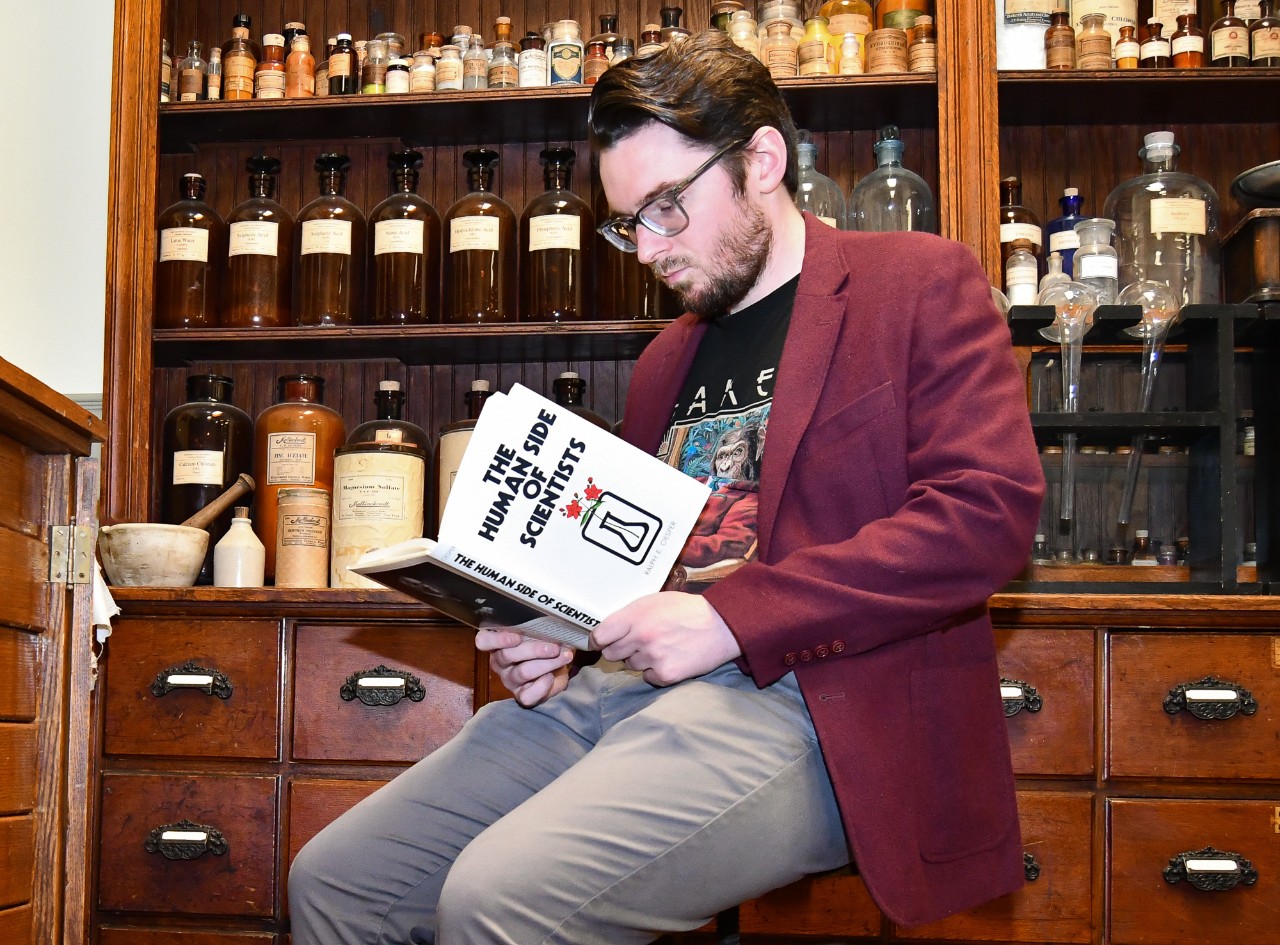 Mark Chalmers reads a book titled The Human Side of Scientists while sitting on a stool surrounded by antique chemistry equipment and glass bottles.