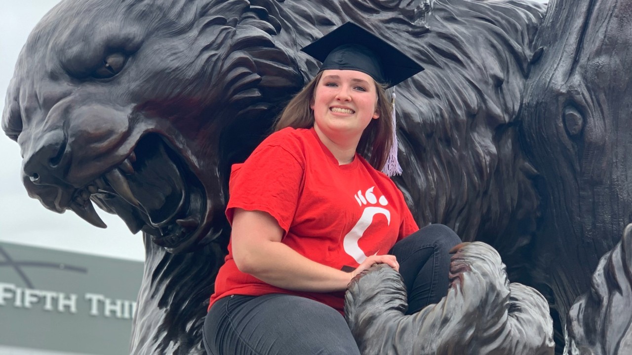Maddie Bell poses with a grad cap on while sitting on the Bearcat statue