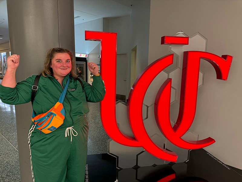 Jac Kern forms a "U" with her arms near a University of Cincinnati logo