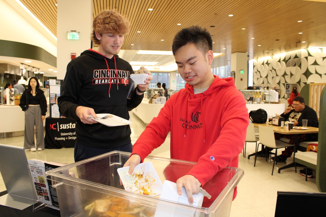 Two male UC students place food waste in plastic container as part of a Weigh the Waste event