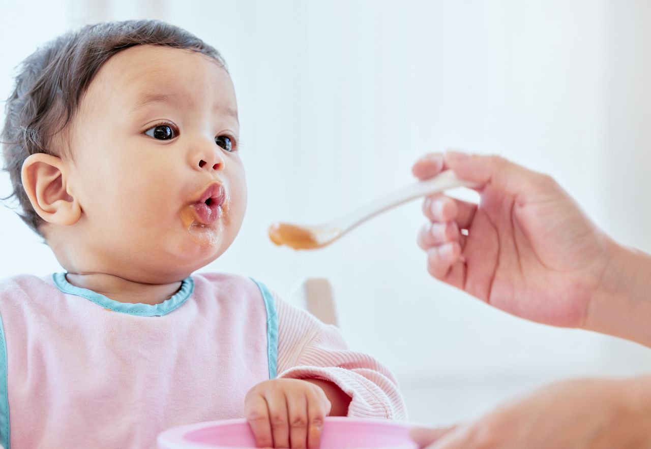 Baby sitting in a highchair and being spoon fed orange pudding