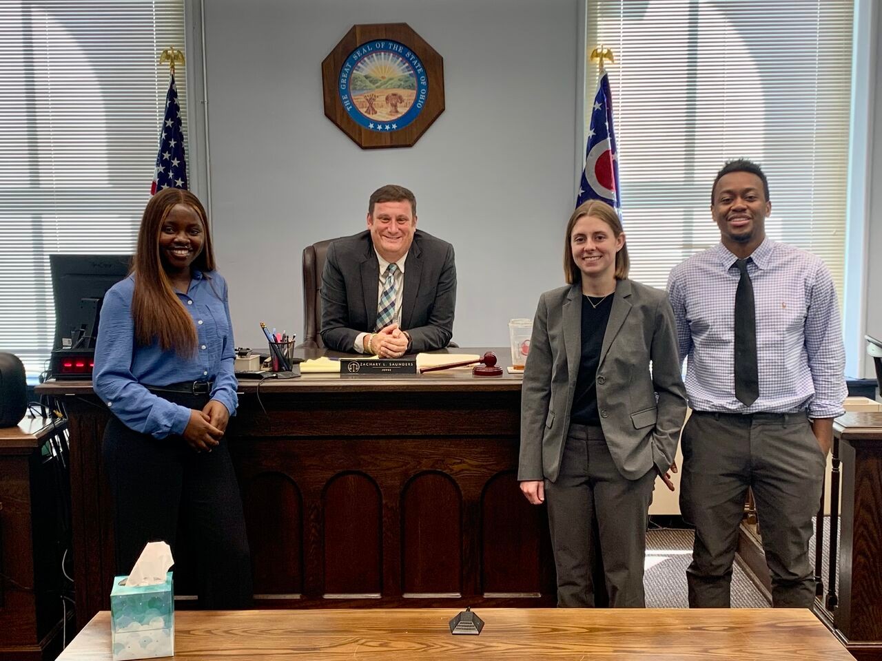 Adesewa Adeyefa is standing while Judge Zachary Saunders is sitting at a desk and Caroline Waller and Kofi Adyepong are standing